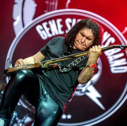 Gene Simmons tocando el bajo con la mano en el mástil, frente al logo de la Gene Simmons Band