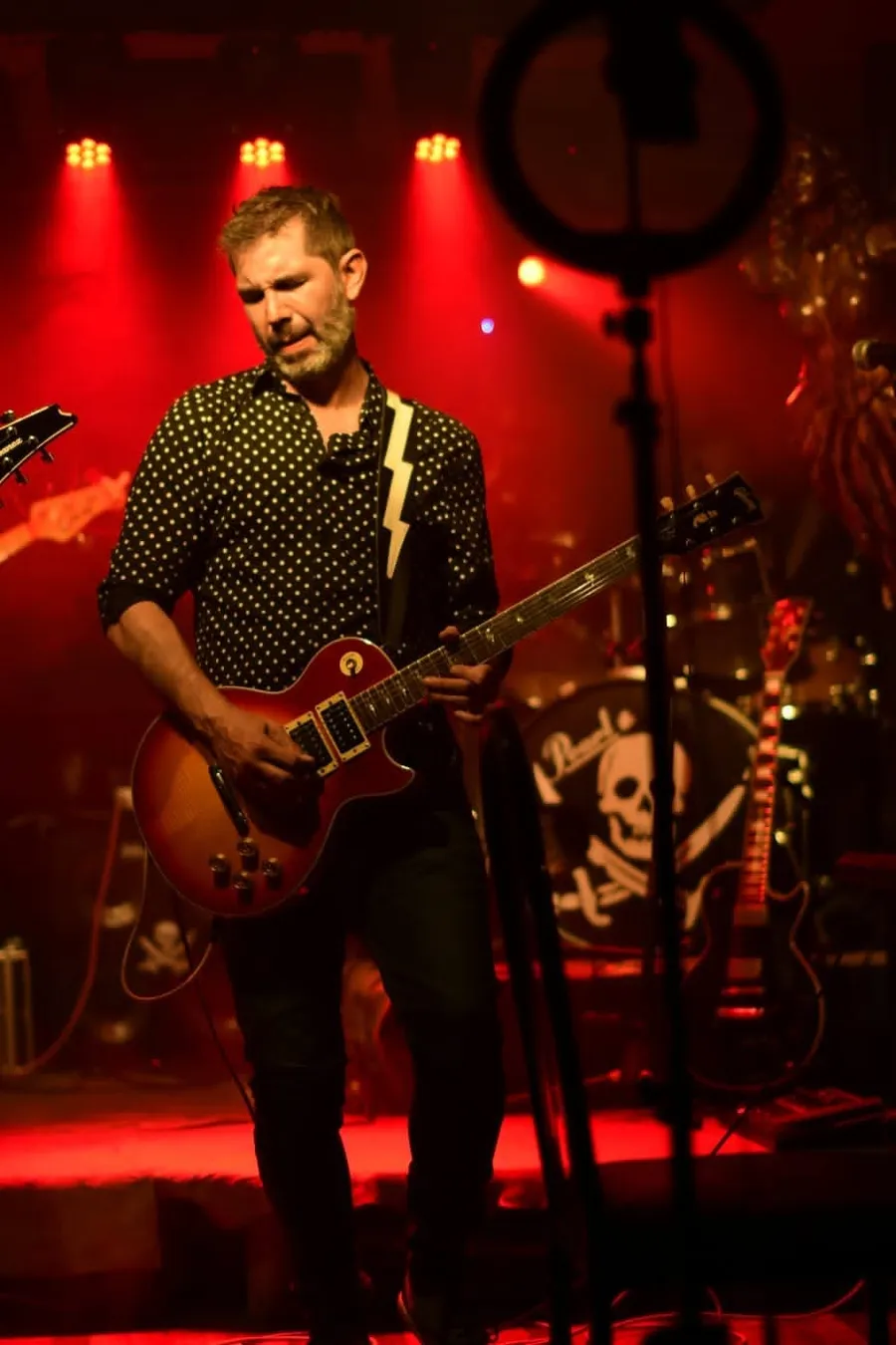 Carlos Sanguinetti tocando la guitarra en vivo, con camisa de lunares y luces rojas de fondo